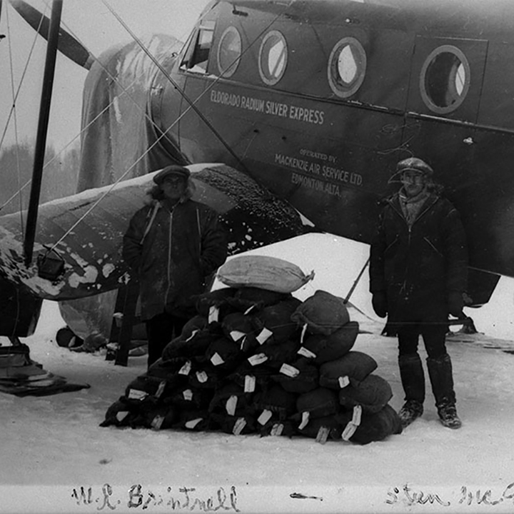 Two men stand in the
snow in front of an airplane that has small round windows and a propeller.
Between the 2 men is a stacked pile of uranium concentrate.