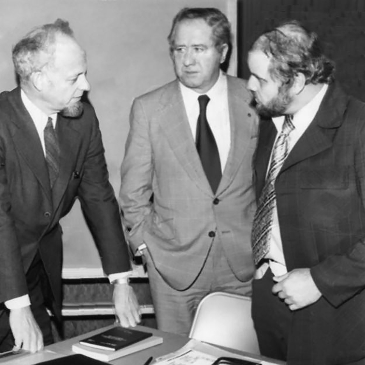 Standing around a
table, a man leans on the table with both hands, while 2 other men are
listening. All the men are in suits, and on the table are a few books and
report binders. 