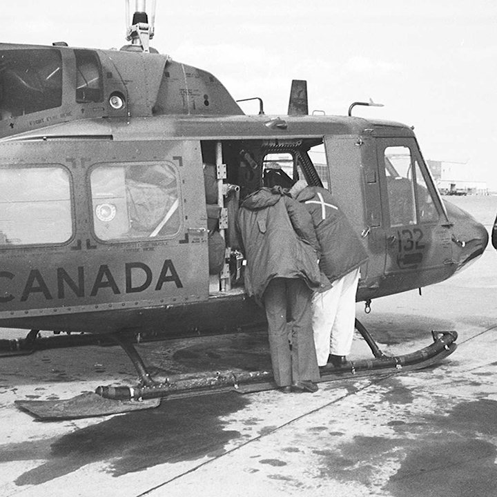 A Canadian Armed
Forces helicopter is landed on the ground. At the open side door, 2 men
lean in. A third man is walking in front of the helicopter.