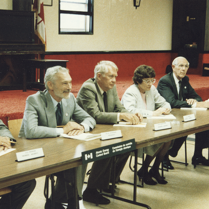 Avec un piano droit
sur une estrade en arrière-plan, 5 membres de la Commission de la CCEA
sont assis à une table et parlent aux membres du public. Une affichette
pliée portant le nom du membre est posée devant chacun deux.