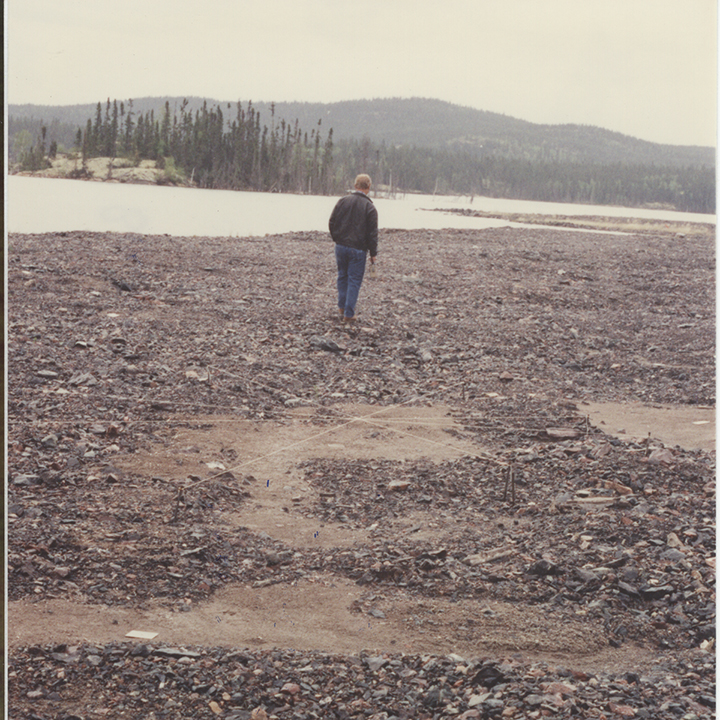 An employee
stands alone on a beach scattered with rocks and pieces of wood. A small
body of water and the treeline on the opposite shore are in the background.
There is a mountain in the distance.