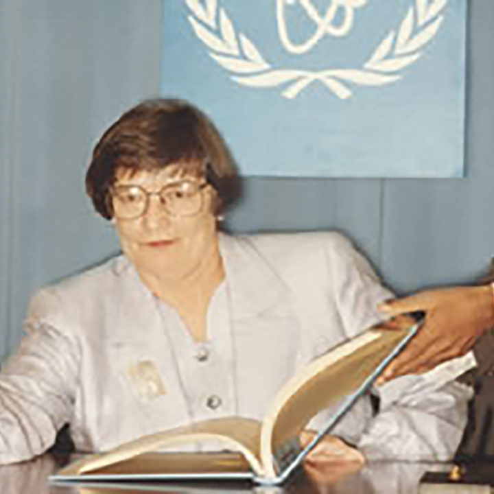 A man holds a large
book open for the Atomic Energy Control Board President to sign. She is
seated at a desk and holds a fountain pen, ready to sign.