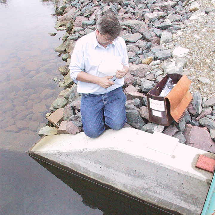 An employee kneels
on a concrete edge to look at a water sample. Beside him is a leather case
with supplies and equipment. The water is still and a rock shore extends
behind him. Trees are reflected in the water. 