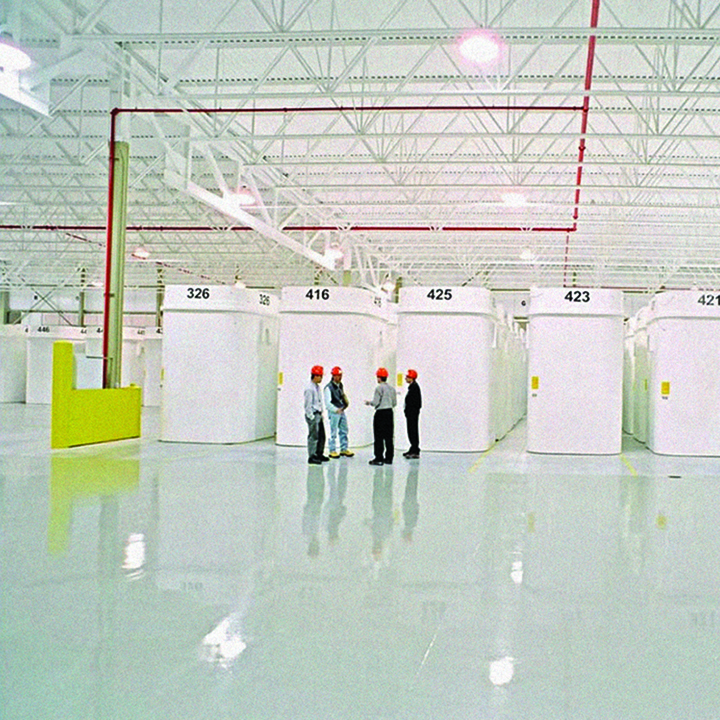 In an expansive white industrial space, 4 employees wearing hard hats
stand in front of several very large containers. The floor is so clean that
it shines, reflecting the light from the ceiling. 