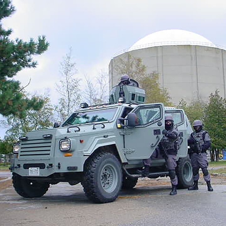 Members of a security response team enter a light armoured vehicle
during a security exercise. The door is open and 1 person is getting into
the vehicle. In the background, there is a large round cement building
surrounded by trees.