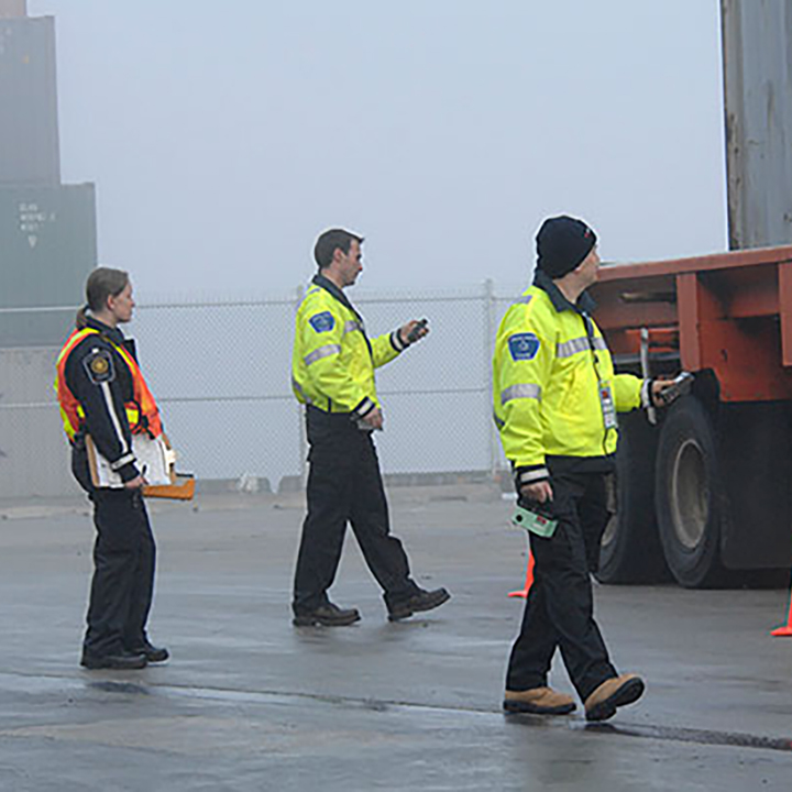 On a foggy day, 3
CNSC employees approach a transport truck surrounded by orange pylons. Two
are wearing identical bright yellow jackets. One wears a reflective vest
and carries a clipboard. 