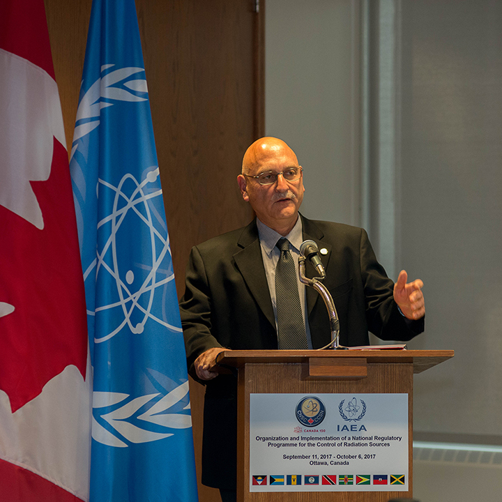 Standing at a podium,
in front of the Canadian flag and IAEA flag, the speaker raises his hand
for effect. On the podium face is a sign identifying the conference.