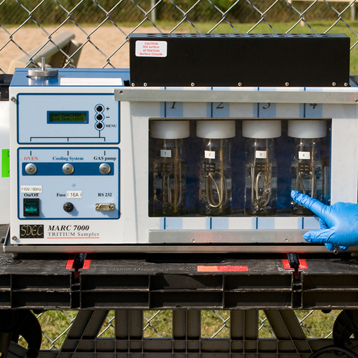 A CNSC employee measures tritium levels using a large piece of
equipment placed on a portable table, which is set up outside against a
chain link fence. She is wearing protective gloves and pointing to a tube
visible through the glass face of the equipment.