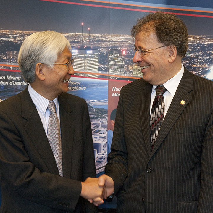 Two men in dark suits
smile and shake hands in front of a colourful backdrop.