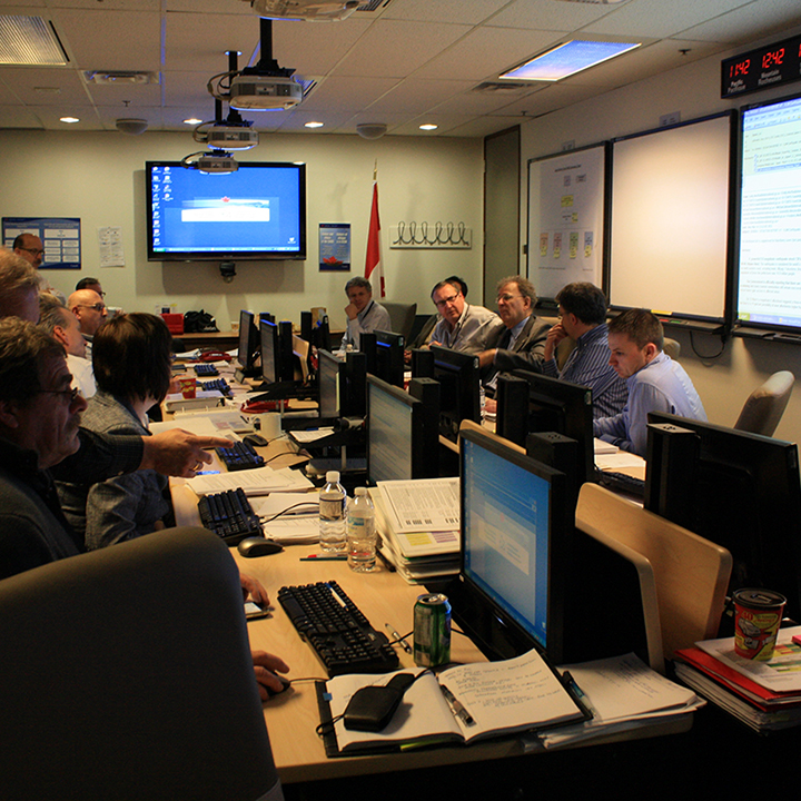 A room crowded with
people, with some seated around a large boardroom table and others
standing. There are many computers and binders on the table. Large monitors
and whiteboards hang around the room on the walls. A Canadian flag stands
in the corner.