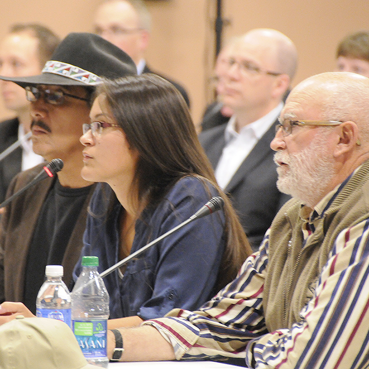 An
indigenous woman speaks into the microphone at a Commission hearing. Seated
on either side of her are 2 other First Nations representatives. In the
background, audience members are seated.