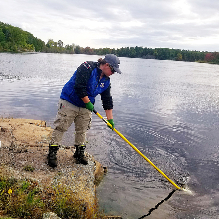 An employee standing
on a rock reaches into the water with a long tool to collect a sample,
leaving ripples in the water. Behind her onshore are 2 plastic containers.

