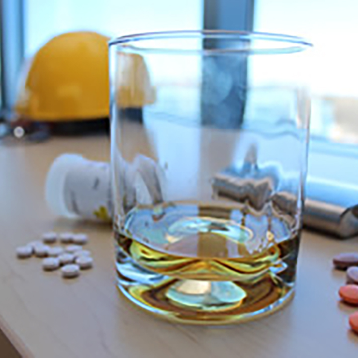 On a desk are a glass
of alcohol, a pill bottle, scattered pills and a vape to denote prohibited
materials. A hardhat is in the background to represent the concept of an
employee's fitness for duty.
