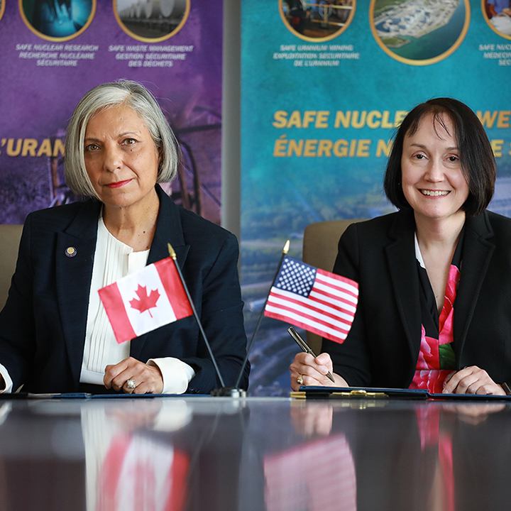 La présidente de la
CCSN et la présidente de la NRC des États-Unis tiennent un stylo et se
préparent à signer un accord. Des petits drapeaux du Canada et des
États-Unis sont posés sur la table entre elles. Des affiches colorées
sont derrière elles.
