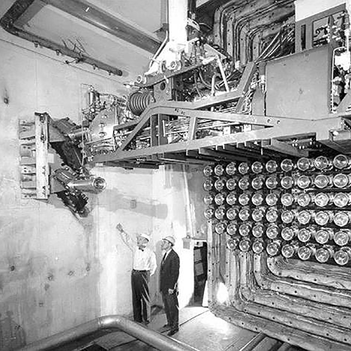 Two workers stand in
front of the Nuclear Power Demonstration reactor. Behind the workers is a
series of tubes. Above the workers, a unit protrudes from the wall. One man
is pointing at the equipment while the other man, in a suit and hard hat,
listens.