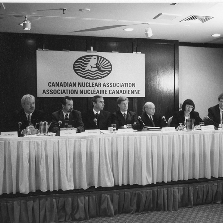 Panel de 8 personnes
assises à une table dhonneur surélevée. Une bannière de lAssociation
nucléaire canadienne est accrochée derrière elles.