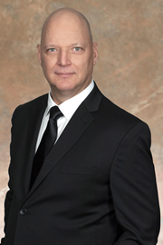 Portrait de Timothy Berube, Ph. D., commissaire, vêtu d’un complet foncé, d’une chemise blanche et d’une cravate, sur un arrière-plan brun clair.