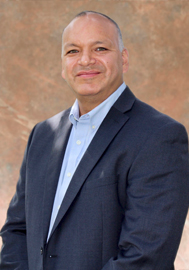 Portrait de Randall Kahgee, commissaire, vêtu d’un complet gris foncé et d’une chemise bleu clair, sur un arrière-plan brun clair.