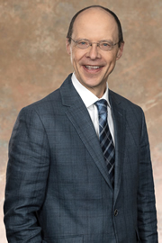 Portrait de Marcel Lacroix, Ph. D., commissaire, vêtu d’un complet gris, d’une chemise blanche et d’une cravate, sur un arrière-plan brun clair.