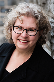 Headshot of Commission member Dr. Victoria Remenda, appearing in a black suit jacket and top against a blurred brown background.