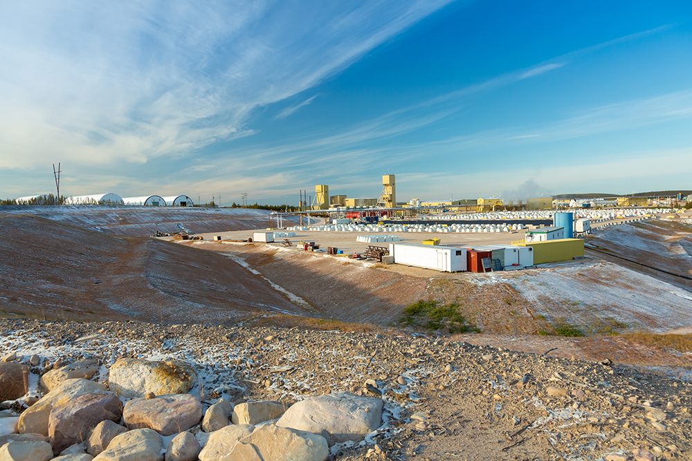 In the bottom left corner is a small cluster of beige and yellow-toned rocks that leads to a downward slope and then up slightly to a large, flat, open area. The ground is made of reddish-brown dirt that is partially covered by light snow and patches of grass. In the middle of the open area are a few white, red and yellow rectangular crates. Behind the crates are many small, white, square structures lined up in rows. In the distance are tall yellow buildings to the left and short, wide ones to the right. A small smoke cloud comes from the right-side buildings. The sky is mainly blue but is slightly overcast towards the horizon.