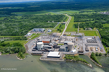 To the left, rear and right of the facilities are green fields of grass and wooded areas. The fields are separated by paths and a road leading to a paved area with a parking lot and multiple beige, orange and black buildings that vary in size. Two large cylinder-shaped buildings stand on opposite sides of the paved area. The buildings are situated on the edge of a waterfront.