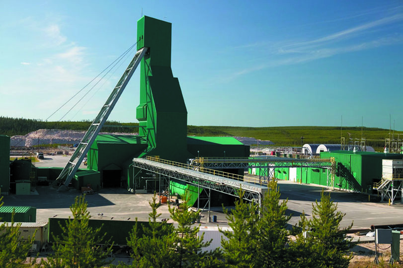 A rectangular green building is in the middle of a paved lot surrounded by grass and trees. There is a tall, slim portion of the building that extends into the sky. Attached to its side is a metal ladder-like structure that angles out to the ground. Surrounding the building are various metal structures and crates.