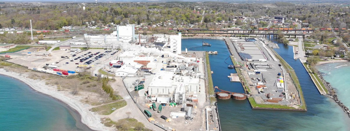 The facility is made up of multiple white buildings that sit on a large, paved lot. The lot leads down to a body of water with a sand-and grass-covered shore. To the right of the buildings is a harbour with boats. A small, rectangular piece of paved land splits the harbour in two down the middle. Behind is a larger land mass populated with trees and buildings.