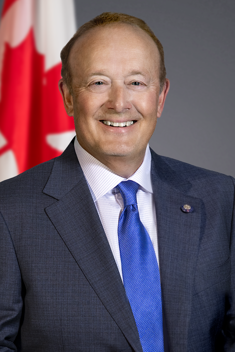 A headshot of Pierre Tremblay, smiling. He wears a dark suit jacket, white shirt and a dark spotted tie. The background is grey.