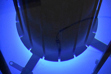 Photo of the SLOWPOKE-2 in-pool-type reactor at the Royal Military College of Canada in Kingston, Ontario. A long black cylinder is vertically submerged in a dark pool of water. A bright light emanating from the bottom of the cylinder lights up the pool in blue. There are 3 rectangular metal bars protruding horizontally from the bottom of the cylinder.
