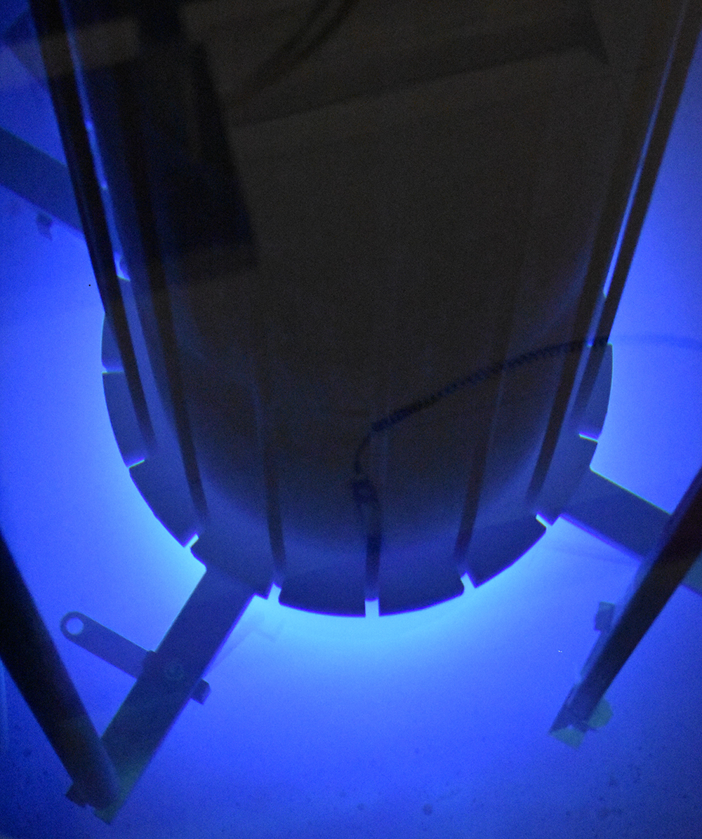 Un grand cylindre noir est immergé verticalement dans une piscine sombre. Une lumière vive émanant du fond du cylindre illumine l’eau en bleu. Trois barres métalliques de forme rectangulaire dépassent horizontalement du fond du cylindre.