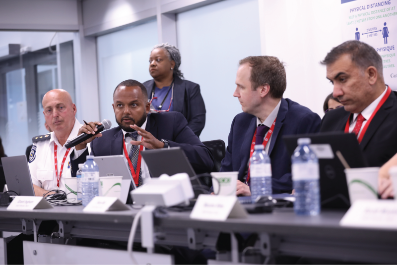 Four people seated at a meeting table. One is speaking into a microphone and gesturing with his left hand.