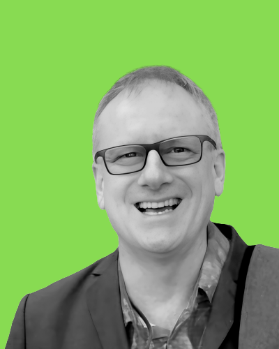 Sur un fond vert vif, un homme avec les cheveux courts et portant des lunettes ainsi qu’une tenue de ville sourit à la caméra.