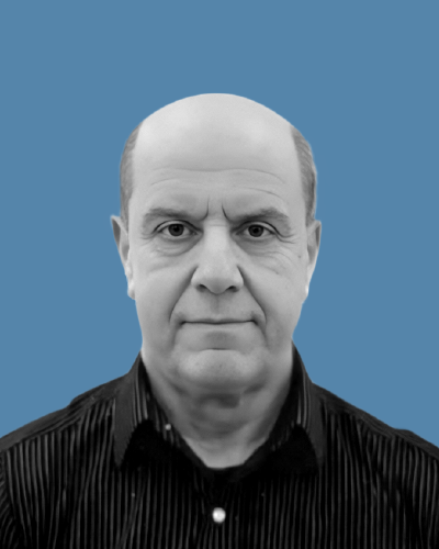 A man with short hair is smiling at the camera.  He is wearing a striped buttoned up shirt. The background is sky blue.